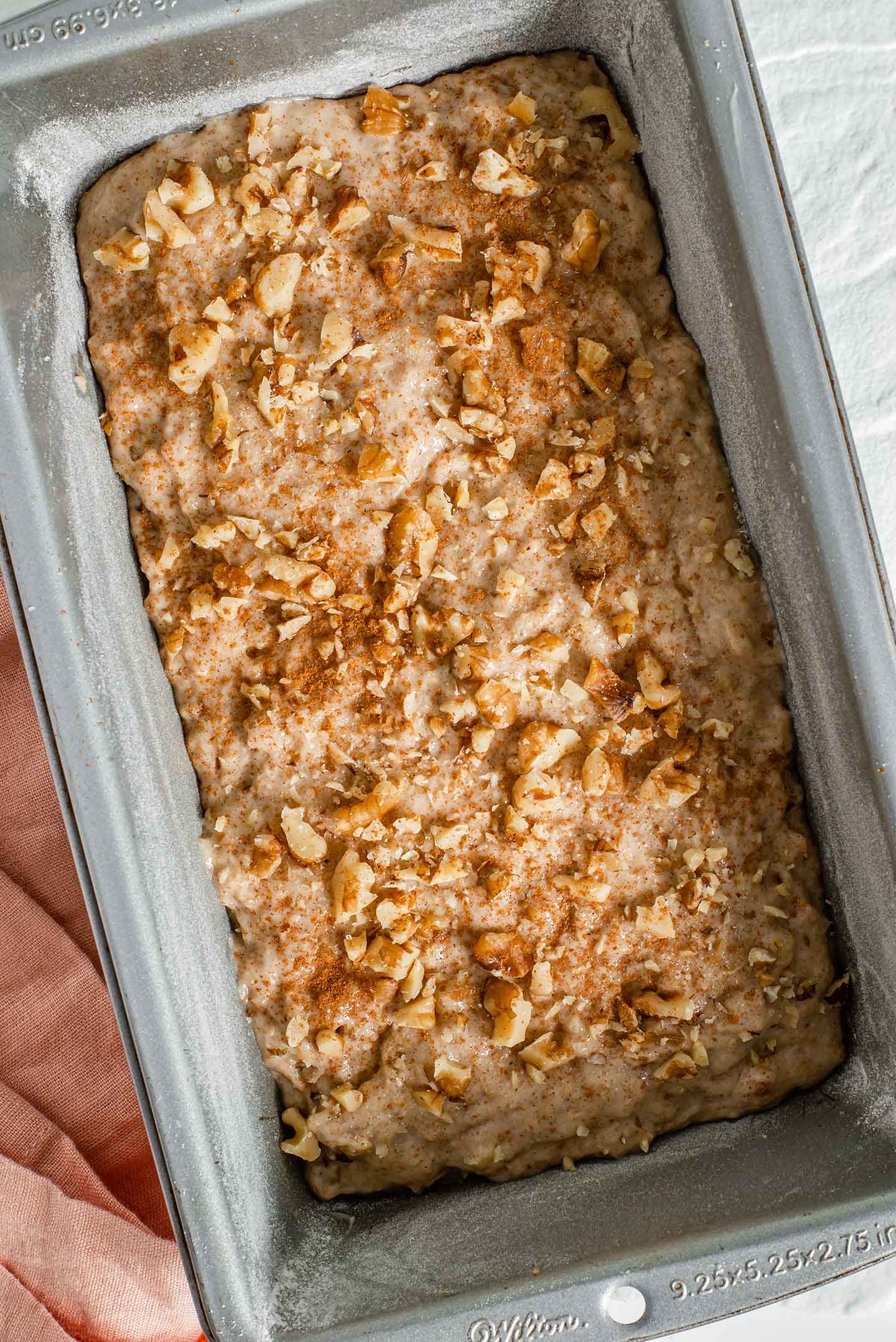 Top down view of vegan banana bread batter poured into a loaf pan. The top of the batter is sprinkled with chopped walnuts, sugar, and cinnamon.