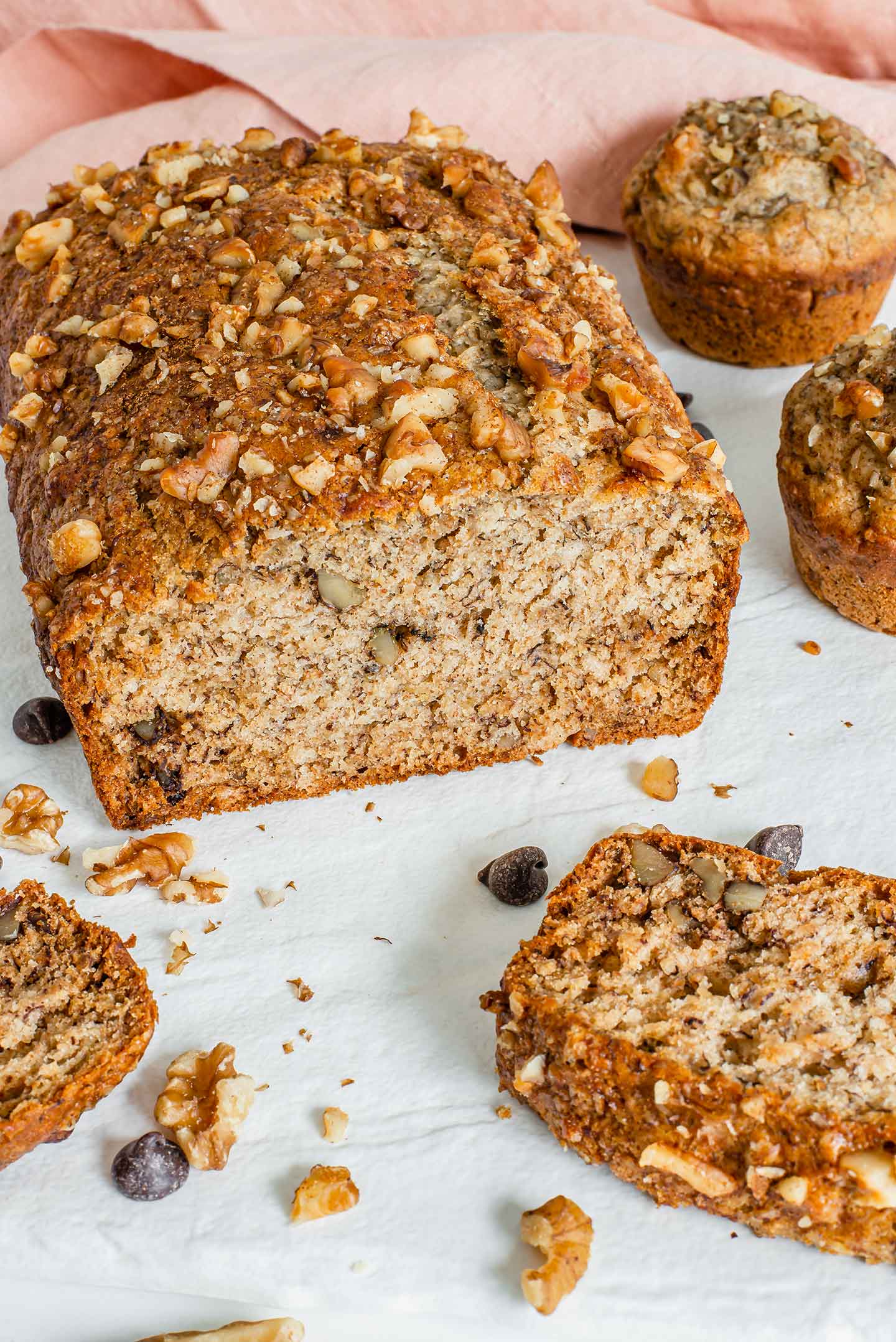 Side view of a sliced loaf of vegan banana bread with walnuts. Banana muffins are visible in the background.