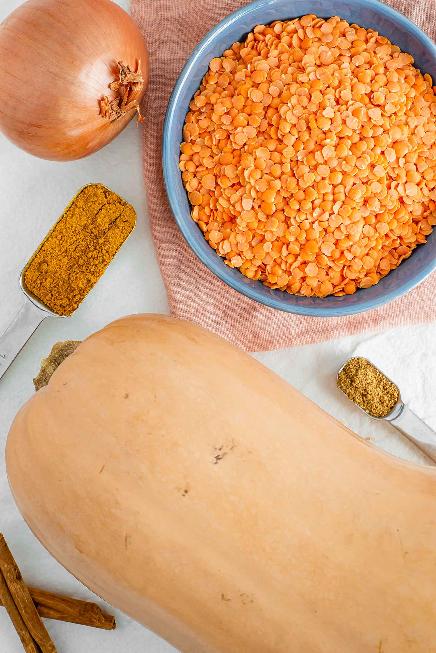 Top down view of a butternut squash, a bowl of dry red lentils, an onion and some seasonings in measuring spoons.