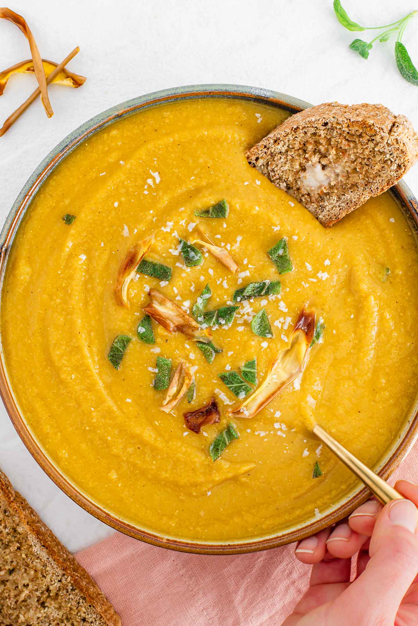 Top down view of a hand dipping a spoon into butternut squash red lentil soup. A buttered piece of whole wheat bread rests in the soup.