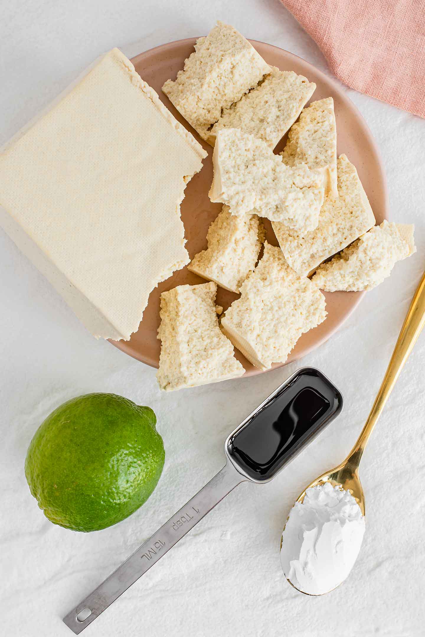 Top down view of a block of extra firm tofu pulled apart into uneven pieces. A fresh lime, tamari, and a spoon of starch rest beside the pieces.