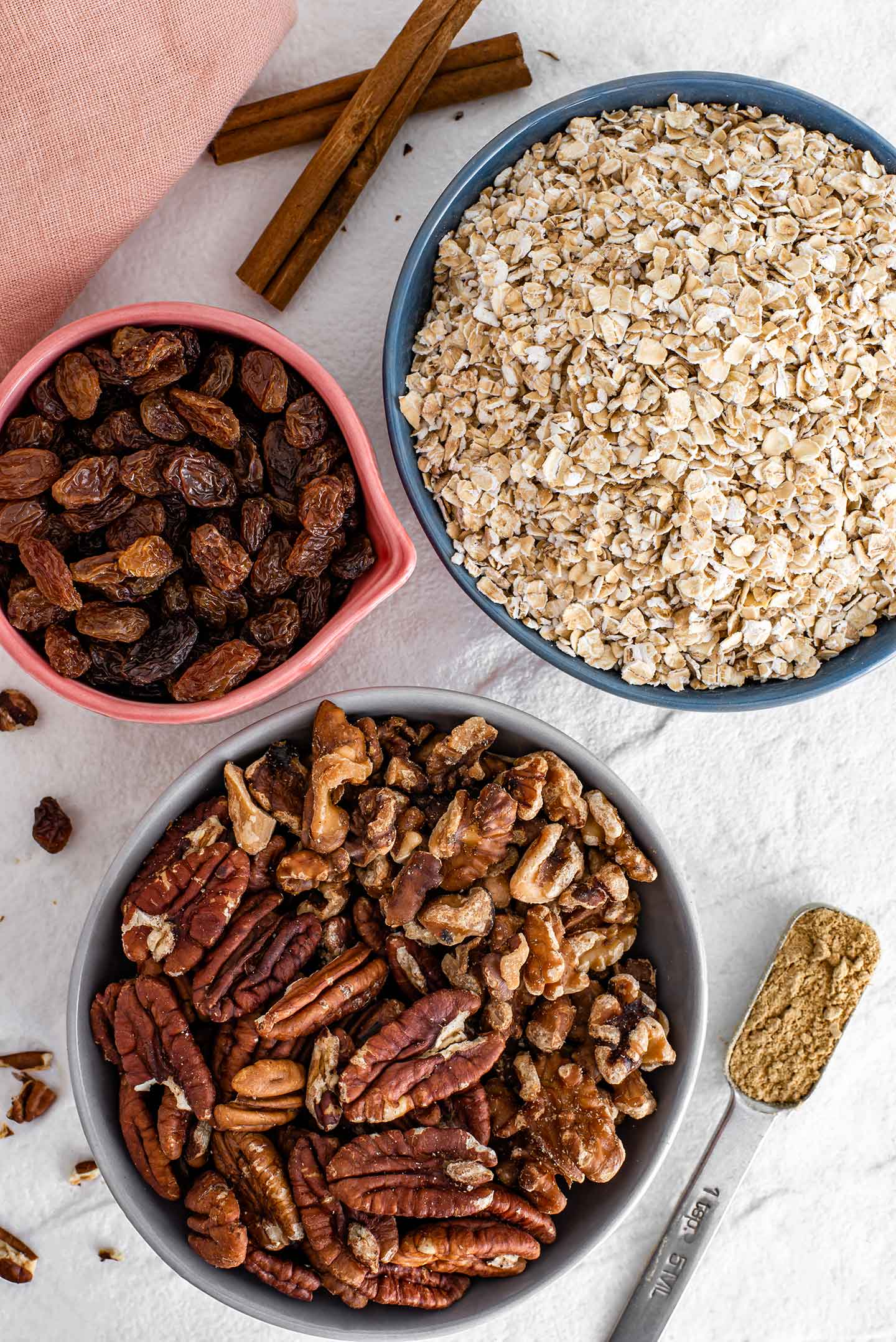 Top down view of ingredients on a white tray. Toasted pecans and walnuts, quick oats, raisins, ground ginger, and cinnamon sticks. 