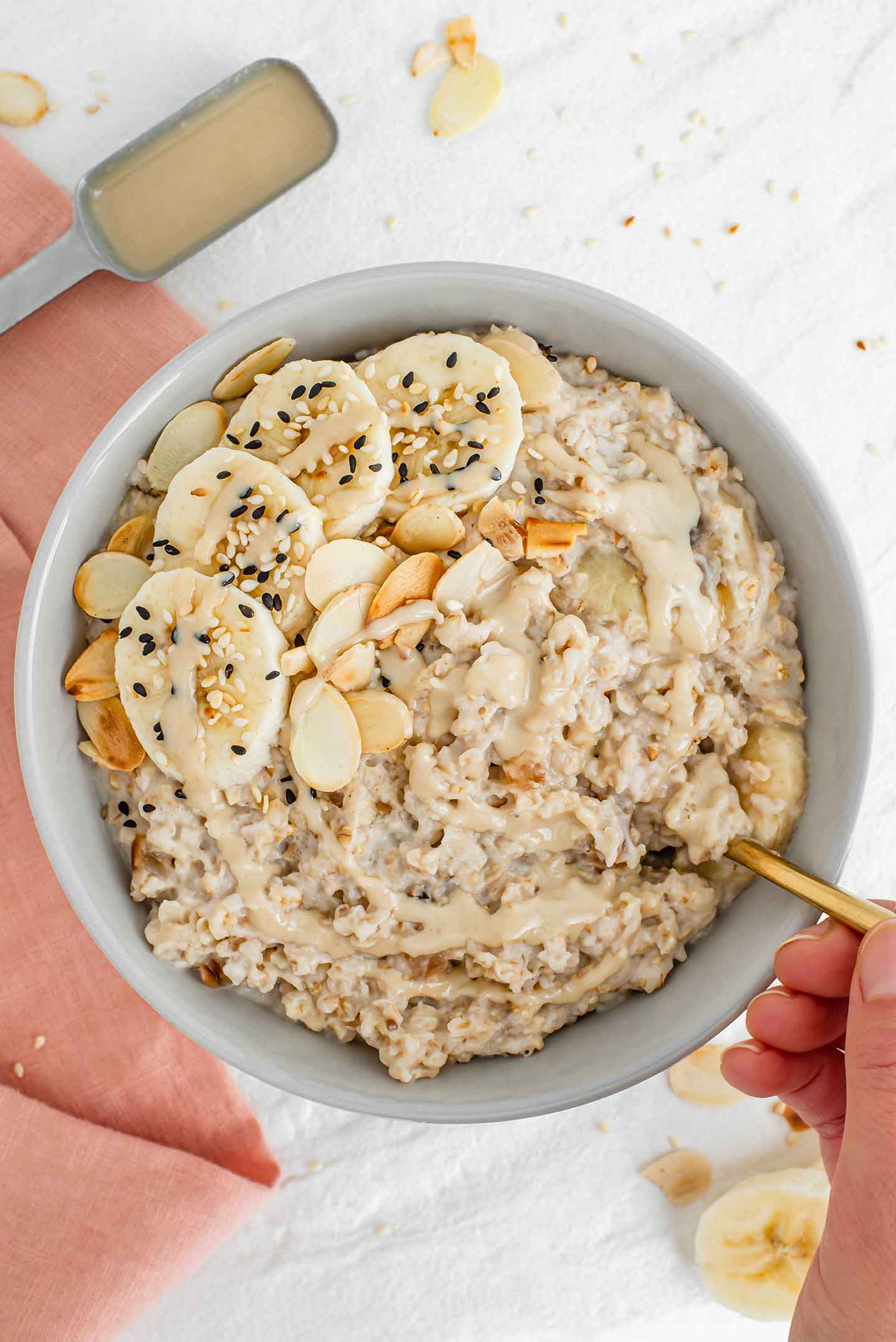 Top down view of a hand dipping a spoon into a bowl of maple tahini oatmeal topped with sliced banana, toasted almonds, toasted sesame seeds and drizzled with extra tahini.
