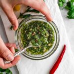 Top down view of cilantro oregano chimichurri being stirred in a small dish with a hot pepper and fresh cilantro to the side.