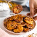 A plate of caramelized plantains. One golden piece is lifted above the plate on a fork.