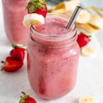 A pink smoothie fills a glass jar. Fresh strawberry and banana slices garnish the side of the glass.