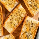 Top down view of golden slices of garlic bread on a wooden tray sprinkled with garlic powder and dried basil.