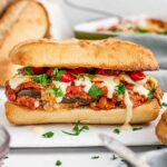 An vegan eggplant parmesan sandwich sits on a white tray. Vegan cheese sauce oozes out of the bun.