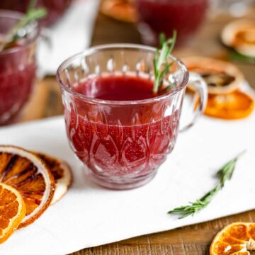 Side view of a small punch glass on a white tray filled with a deep red bubbly punch. A sprig of rosemary garnishes the glass.