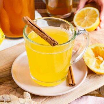 Bright yellow tea in a glass mug is surrounded with fresh ginger, lemon, and garnished with a cinnamon stick.