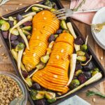 Top down view of a roasted hasselback butternut squash on a tray with parsnips, beets, and brussels sprouts.