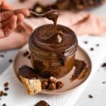 A spoon scoops thick and creamy vegan Nutella hazelnut spread out of a glass jar.