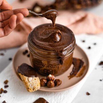 A spoon scoops thick and creamy vegan Nutella hazelnut spread out of a glass jar.