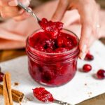 A hand lifts a spoon of cranberry sauce from a jar full of the festive red sauce. Cinnamon sticks and fresh cranberries are scattered around the jar.