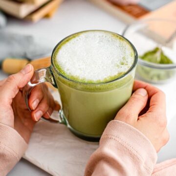 Hands wrap around a green matcha latte in a glass mug topped with white foam.