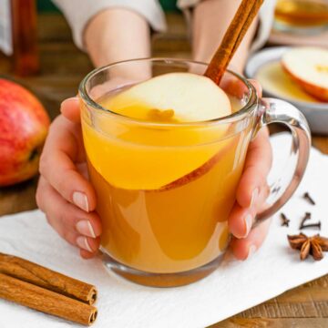 Side view of hands wrapped around a warm glass mug of hot apple cider wassail garnished with an apple slice and cinnamon stick.