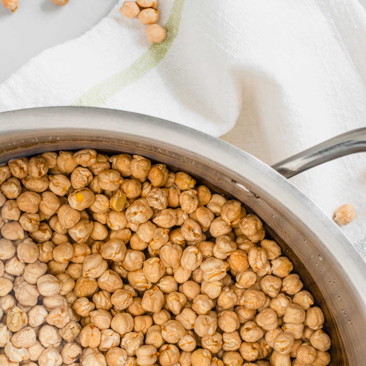Top down view of dried chickpeas in a large pot of water. A dish towel with scattered dried chickpeas lays underneath.