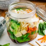 Side view of a glass jar with a clamp lid filled with vegetables, tofu, rice vermicelli noodles, and a dark broth.