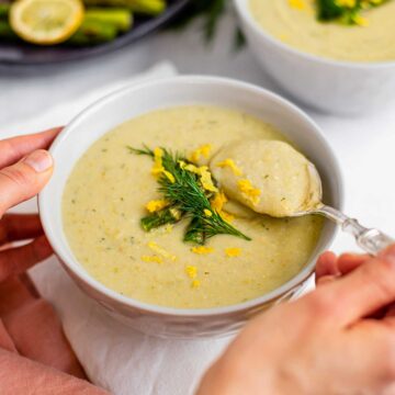 A spoon scoops thick and creamy lemon asparagus soup from a bowl garnished with roasted asparagus spears, fresh dill, and lemon zest.