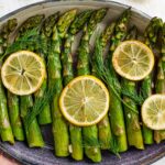 Top down view of roasted asparagus spears in a serving platter with lemon slices and fresh dill to garnish.