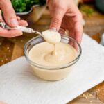 A hand lifts a spoon of creamy lemon tahini dressing from a small bowl. The dressing is thick but pourable.