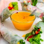 Side view of a small bowl of spicy mango sauce surrounded by fresh rolls with lettuce, mango, and red bell pepper.