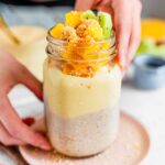 Side view of a colourful jar of mango yogurt overnight oats. Fresh mango, toasted coconut and some slices of kiwi sit on top.