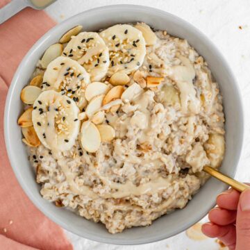 A hand dips a spoon into a bowl of creamy oatmeal topped with sliced banana, toasted almonds and sesame seeds.