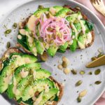 Two slices of miso avocado toast on a plate topped with pickled red onion, seeds, and drizzled with miso glaze.