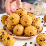 A hand reaches for a chickpea peanut butter energy ball speckled with chocolate chips and displayed in a towering mound of energy balls.