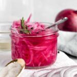 Side view of quick pickled red onions in a glass jar with vinegar, sugar, salt, and garnished with a sprig of rosemary and whole black pepper.