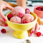 Three scoops of raspberry nice cream garnished with frozen raspberries in a bright yellow sorbet bowl.