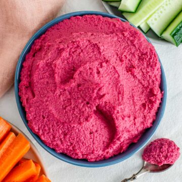 A bowl filled with bright pink beet hummus. Carrots and celery sticks surround the bowl.