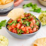 Salsa fresca fills a small dish. A tortilla chip is placed inside the bowl of chunky salsa garnished with cilantro.