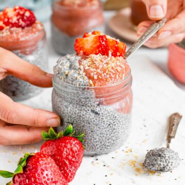 A spoon rests in a small glass jar of creamy chia pudding served with strawberry jam, sprinkled with toasted coconut and garnished with fresh strawberry slices.