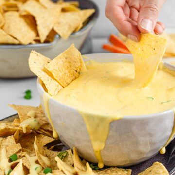 A hand dips a tortilla chip into a large bowl of vegan queso. The queso is thick, creamy, and "cheese" like.
