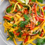 A colourful mango salad with red pepper, red onion, cilantro, mint, crushed peanuts, and lime wedges.