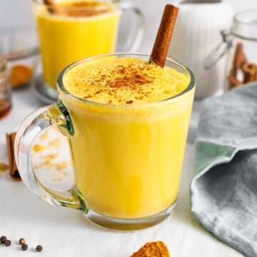 Turmeric golden milk fills a clear glass mug. A cinnamon stick garnishes.