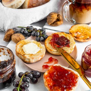 Side view of toasted Portuguese sweet muffins buttered and smeared with jam. Milk is poured into coffee in the background, a bread basket overflows with muffins and grapes and figs surround the cut muffins.