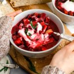 Hands wrap around a big bowl of vegan borscht. Sour cream and dill garnish the hearty soup.