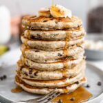 Side view of a towering stack of chia chip pancakes. Maple syrup streams down the sides.