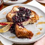 Two stacked golden slices of french toast topped with a blueberry sauce, toasted sliced almonds, and drizzled with maple syrup.