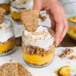 A hand places a personal sized vegan pumpkin pie parfait on a white tray. Layers of pumpkin mousse and granola are topped with whipped cream and a cookie.
