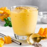 A thick and creamy mango lassi in a glass with mango cubes and mint leaves in the background.