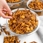 A hand grabs party mix from a large bowl of spiced pretzels and cereals.
