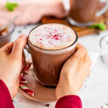Side view of hands wrapped around a glass mug of vegan peppermint hot chocolate garnished with whipped cream and candy cane shavings.