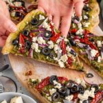 Top down view of a hand lifting a slice of thin crust vegan pesto pizza with feta crumbles from a wooden tray.