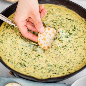 A hand dips a cracker into the centre of a cast iron skillet filled with creamy spinach artichoke white bean dip.