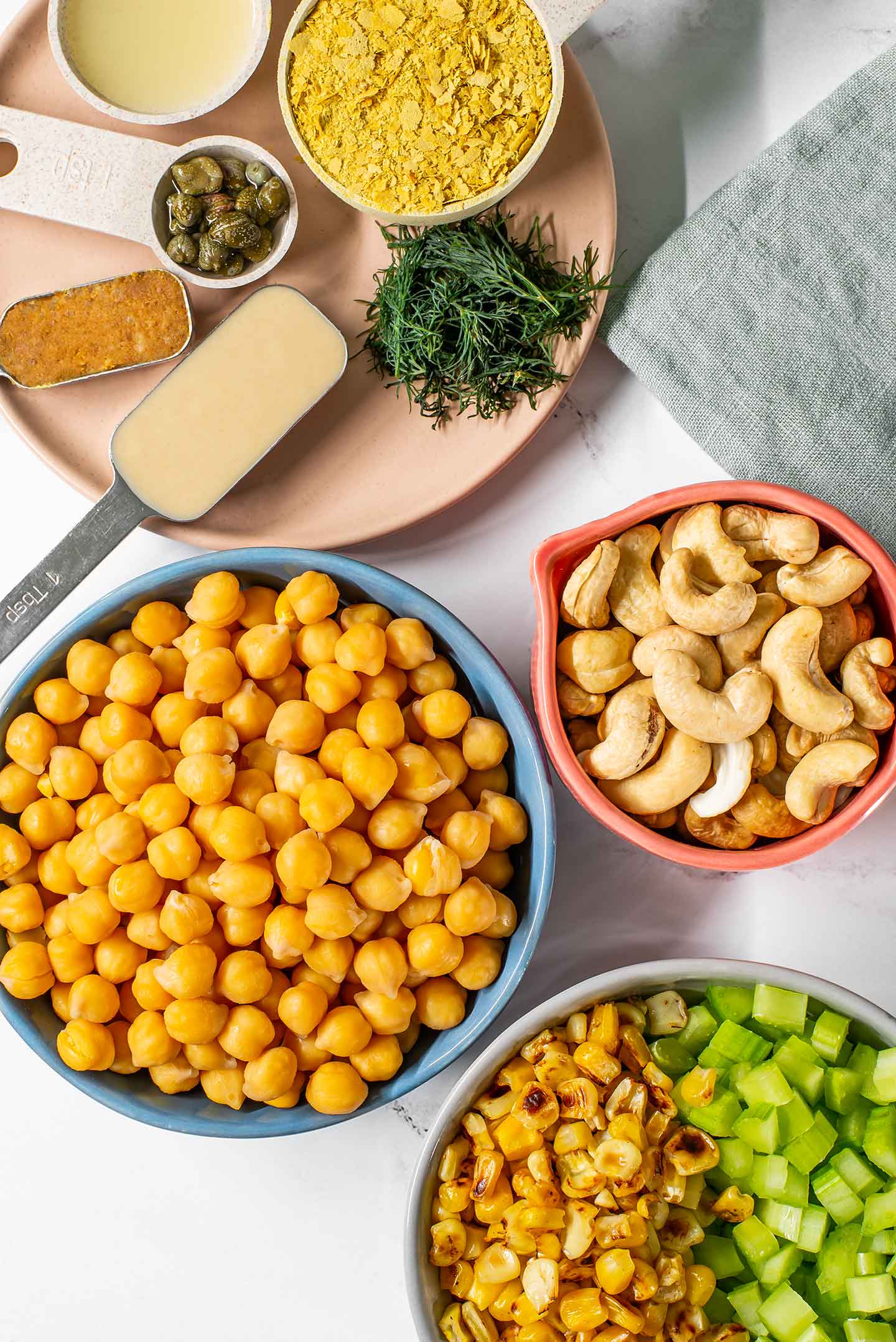 Top down view of ingredients on a white tray. Chickpeas, cashews, charred corn, celery, and a plate of seasonings.