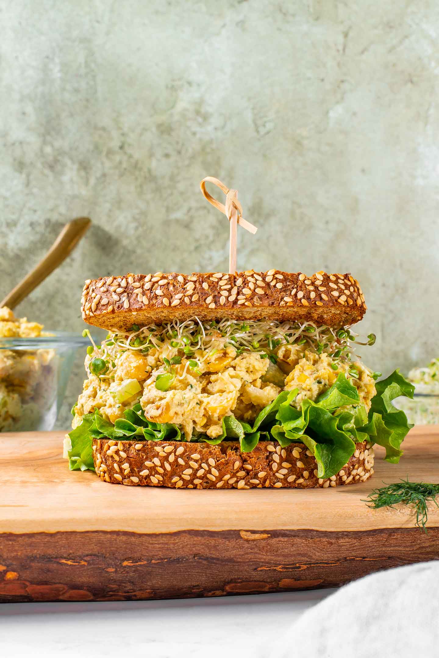 Side view of chickpea smash on lettuce and topped with sprouts between two pieces of seeded bread. A wooden pick holds the sandwich together.